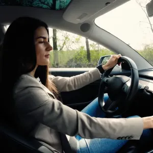 Une jeune femme aux cheveux longs conduit une voiture connectée, son regard tourné vers le tableau de bord numérique.