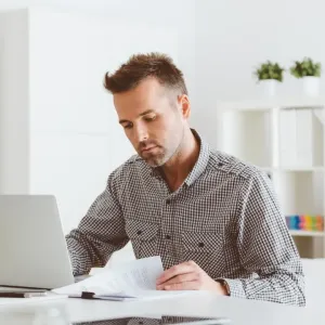 Un homme concentré travaille sur un ordinateur portable, tenant des documents dans un bureau lumineux et épuré.