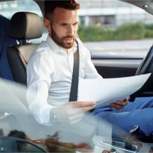 Un homme barbu en chemise lit des documents au volant d'une voiture, un costume à l'arrière.