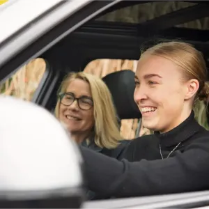 Une jeune femme dans une voiture, mains sur le volant, avec sa mère côté passager.