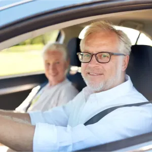 Un homme senior souriant conduit une voiture, sa passagère également souriante à l'arrière-plan.