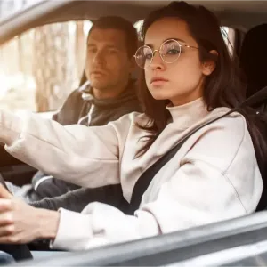 Une femme aux cheveux foncés et lunettes conduit une voiture, regardant attentivement devant elle. Un homme est visible côté passager.