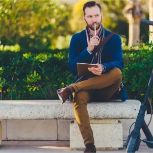 Un homme barbu assis sur un banc, pensif, avec sa trottinette électrique noire garée à côté de lui.