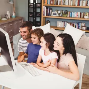 Une famille joyeuse (mère, père, deux enfants) est assise devant un ordinateur de bureau dans un salon lumineux.