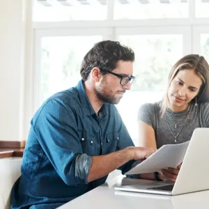 Un couple discute de documents (financiers ou immobiliers) devant un ordinateur portable dans leur cuisine lumineuse.