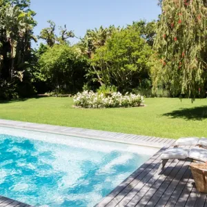 Une piscine rectangulaire avec transats et terrasse, un grand jardin ensoleillé à l'arrière-plan.