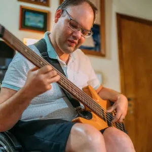Homme en fauteuil roulant jouant de la guitare basse électrique (couleur bois) chez lui, amplificateur visible derrière lui.