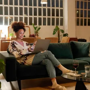 Une femme souriante est assise sur un canapé vert la nuit, utilisant son ordinateur portable. Ambiance chaleureuse avec plantes.