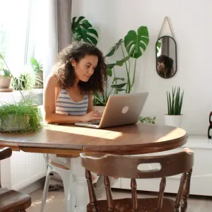 Jeune femme aux cheveux bouclés travaillant sur son ordinateur portable à une table dans une pièce lumineuse remplie de plantes.