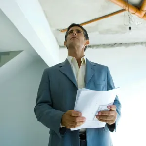 Homme en costume regardant un plafond avec des tuyaux exposés et tenant des plans. Inspection de travaux.