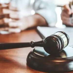 Un gros plan sur un marteau de juge (maillet) posé sur un socle en bois, en face de personnes signant un document juridique.