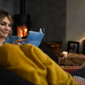 Une femme est détendue sous une couverture jaune, lisant un livre dans un salon sombre et confortable avec un poêle à bois allumé.