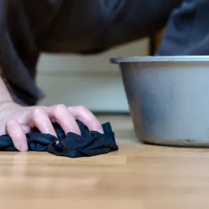 Gros plan sur une main nettoyant le sol en bois avec un chiffon noir, près d'une bassine d'eau.