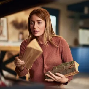 Femme aux cheveux blonds alimentant une cheminée ou un poêle avec des bûches dans un intérieur moderne et chaleureux.