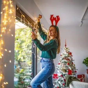 Une jeune femme joyeuse portant des bois de renne décore une fenêtre avec des guirlandes lumineuses à côté d'un arbre de Noël.