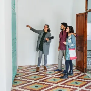 Une agente immobilière montre un détail d'une pièce vide à un couple dans un couloir avec un sol à motifs.
