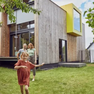 Une petite fille court dans le jardin devant une maison moderne en bois, ses parents en arrière-plan.