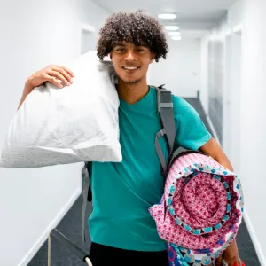 Jeune homme souriant portant un oreiller et un sac de couchage, prêt pour un voyage ou un déménagement.