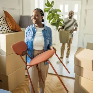Un couple déménage : femme tenant une chaise, homme avec une plante, entourés de cartons dans un intérieur ensoleillé.