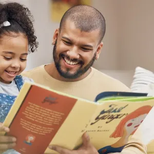 Père et fille souriants lisent ensemble un livre d'histoires sur le canapé. Moment de complicité et d'éducation.