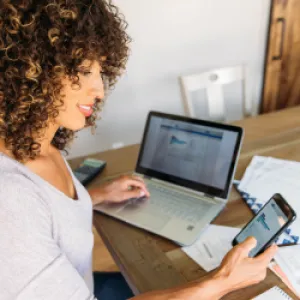 Une femme consulte son smartphone, son ordinateur et des documents papier.
