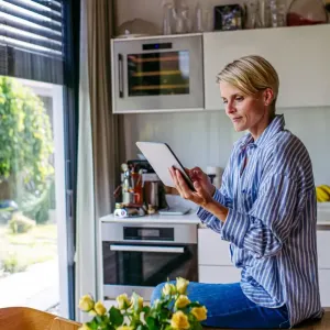 Une femme en chemise rayée utilise une tablette dans une cuisine lumineuse ouverte sur un jardin verdoyant.