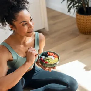Jeune femme aux cheveux bouclés, vêtue d'une tenue de sport verte et noire, assise par terre dans un appartement lumineux et mangeant un smoothie bowl garni de fraises, kiwis, bananes et fruits rouges.