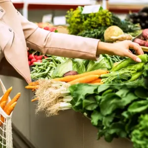 Gros plan sur une personne tenant un sac en filet réutilisable rempli de carottes, tout en choisissant un piment vert sur un étal de marché composé de divers légumes frais et herbes aromatiques.