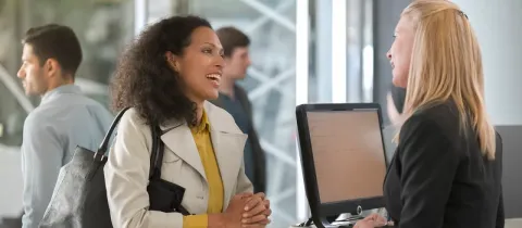 Une femme souriante parle à une employée derrière un comptoir, possiblement à une banque ou réception.