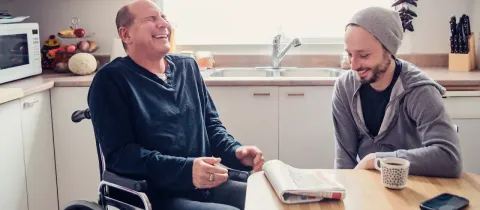 Deux hommes rient ensemble dans une cuisine, l'un en fauteuil roulant et l'autre assis à une table.