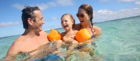 Une famille joyeuse : père, mère et leur fille s'amusent dans l'eau turquoise sous un ciel bleu.
