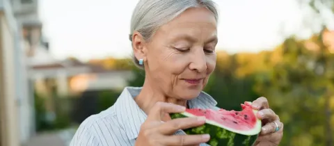Une femme âgée aux cheveux gris sourit en savourant une tranche de pastèque fraîche.