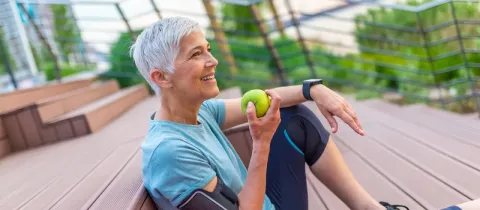 Une femme aux cheveux gris, assise à l'extérieur, sourit en tenant une pomme verte après le sport.
