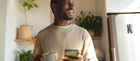 Un homme souriant tient une tasse et un téléphone, regardant au loin dans une cuisine lumineuse.