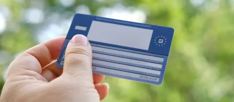 Une main tient une carte européenne d'assurance maladie, sur fond de feuillage vert flou.