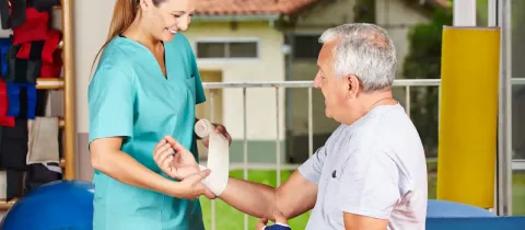 Une physiothérapeute souriante bande le bras d'un homme âgé assis, lui offrant un soutien.