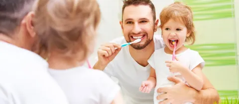 Un père et sa petite fille souriants se brossent les dents ensemble devant un miroir.
