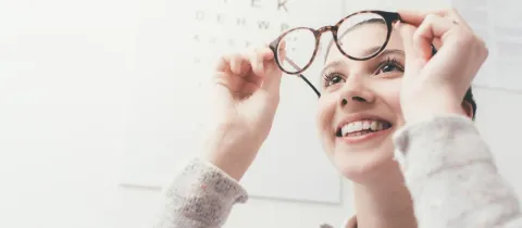 Une jeune femme souriante tient des lunettes devant un tableau optométrique.