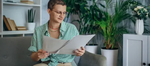 Une jeune femme aux cheveux courts et lunettes lit des documents sur un canapé.