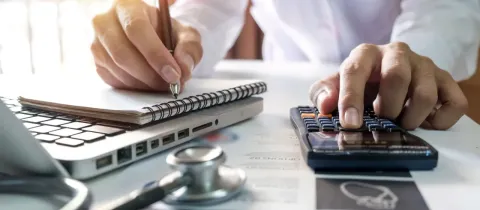 Gros plan sur une personne calculant des chiffres sur une calculatrice, avec un ordinateur portable et un stéthoscope.