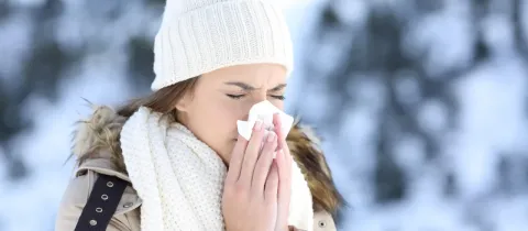 Une femme en tenue d'hiver se mouche dans un mouchoir, l'air souffrant, dans un paysage enneigé.