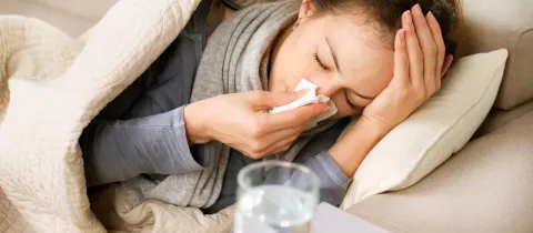 Une femme malade au lit, se mouche dans un mouchoir, avec des médicaments à côté.