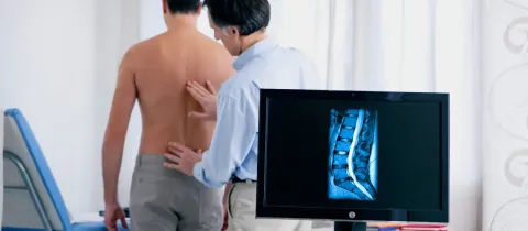 Un médecin examine le dos d'un patient tandis qu'une IRM de colonne vertébrale s'affiche.