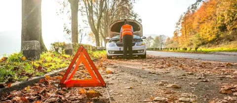 Un homme en gilet de sécurité orange examine le moteur d'une voiture en panne, avec un triangle de signalisation.