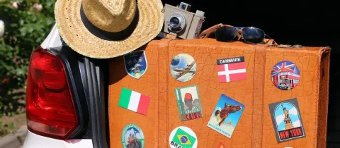 Une valise vintage ornée d'autocollants de voyage est posée à l'arrière d'une voiture, avec un chapeau et une caméra.