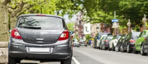 L'arrière d'une voiture grise est garé en épi sur le bord d'une rue bordée d'arbres et d'autres voitures.