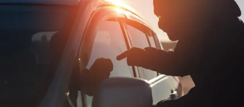 Silhouette d'une personne mal intentionné s'approchant d'une voiture, le soleil couchant créant un fort contre-jour.