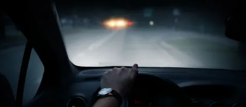 Une main tient le volant d'une voiture, les phares d'un autre véhicule sont visibles de nuit à travers le pare-brise.