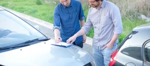 Deux hommes sont penchés sur le capot d'une voiture endommagée, l'un d'eux remplit un constat à l'amiable sur un presse-papiers.