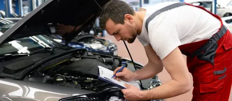 Un mécanicien en salopette rouge et blanche est penché sur le moteur d'une voiture, notant des informations sur un presse-papiers.